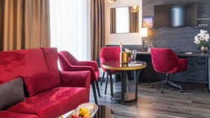 A modern hotel room with a red sofa, matching armchairs, a round dining table with a bottle of champagne and glasses, and a TV mounted on the wall. - PLAZA Premium Grand Winston (Rijswijk)
