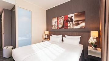 Una habitación de hotel con una cama grande, dos mesitas de noche con lámparas, una obra de arte colgada en la pared y una puerta que da a otra habitación.