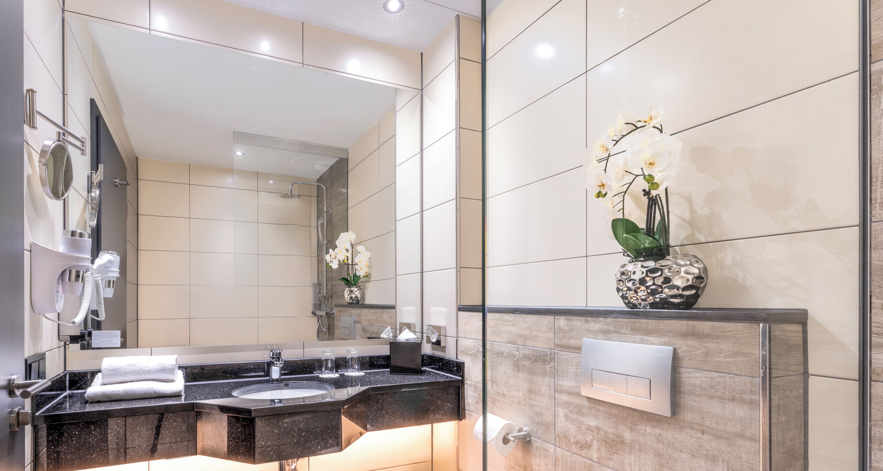 A modern bathroom with a double sink, a large mirror, and a vase with white orchids.
