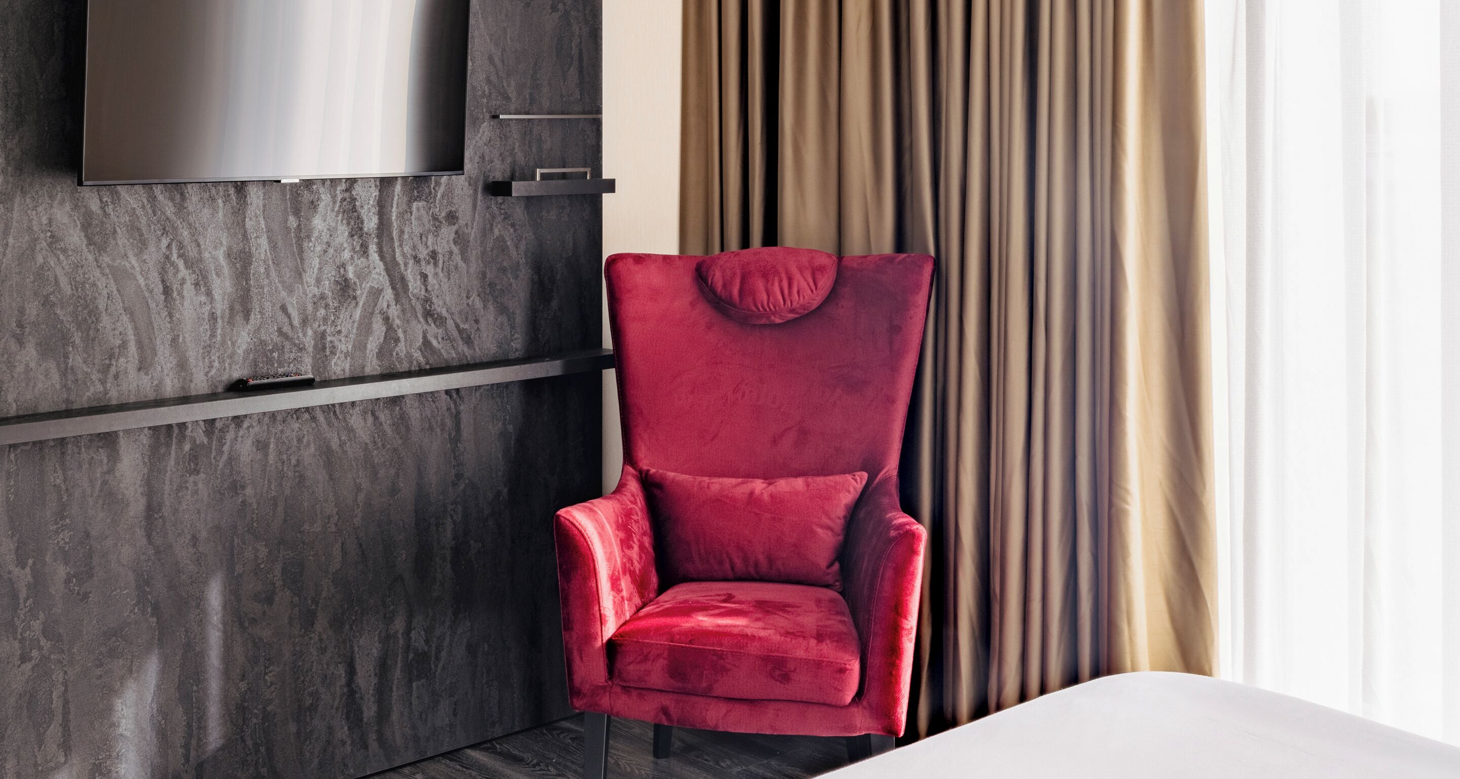 Una habitación moderna con una butaca de terciopelo rojo, un televisor de pantalla plana grande montado en una pared con textura, y vistas a cortinas y una ventana.