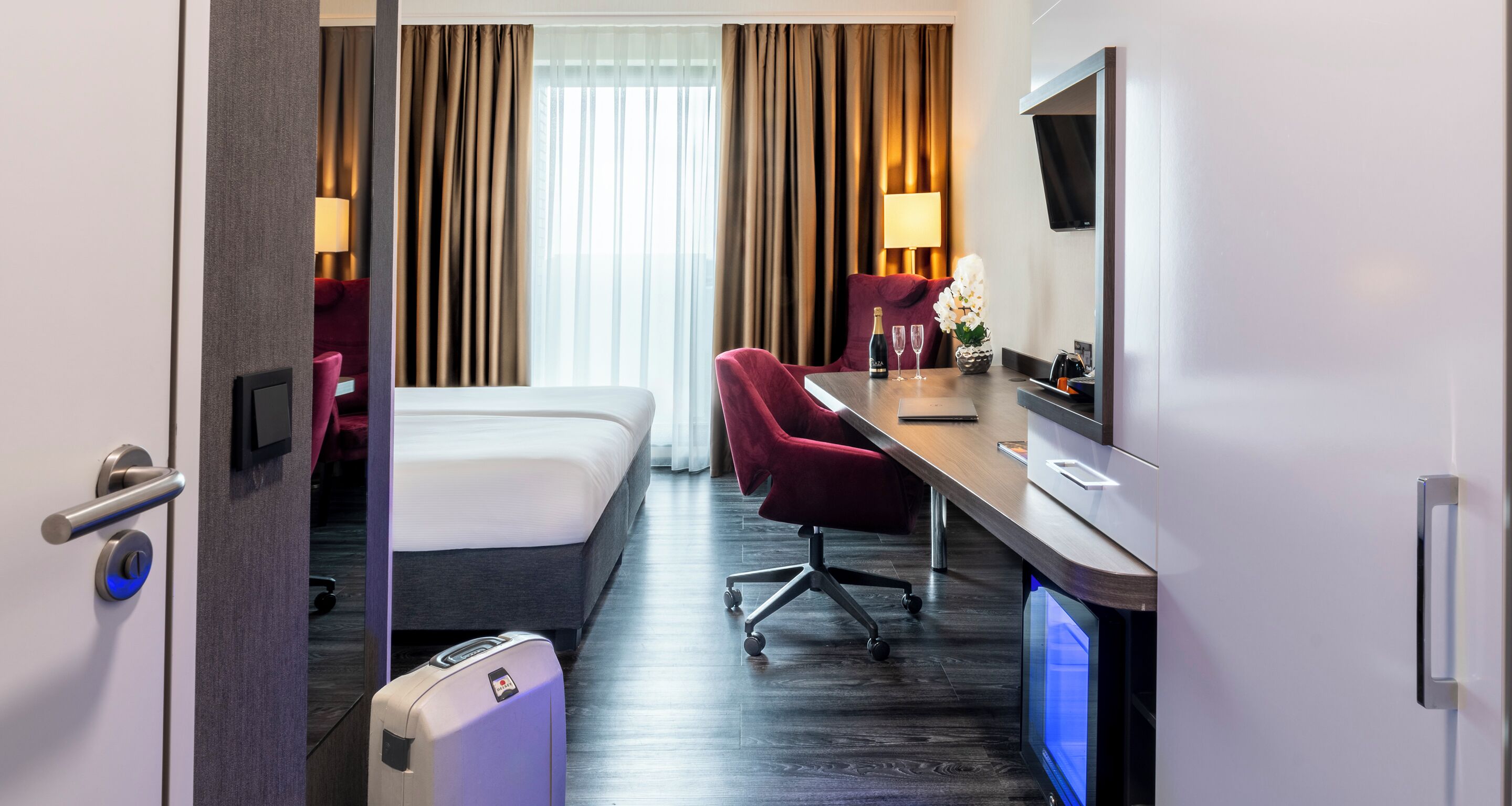 A hotel room with a bed, a desk with a computer, a chair, a TV, and a window with curtains.
