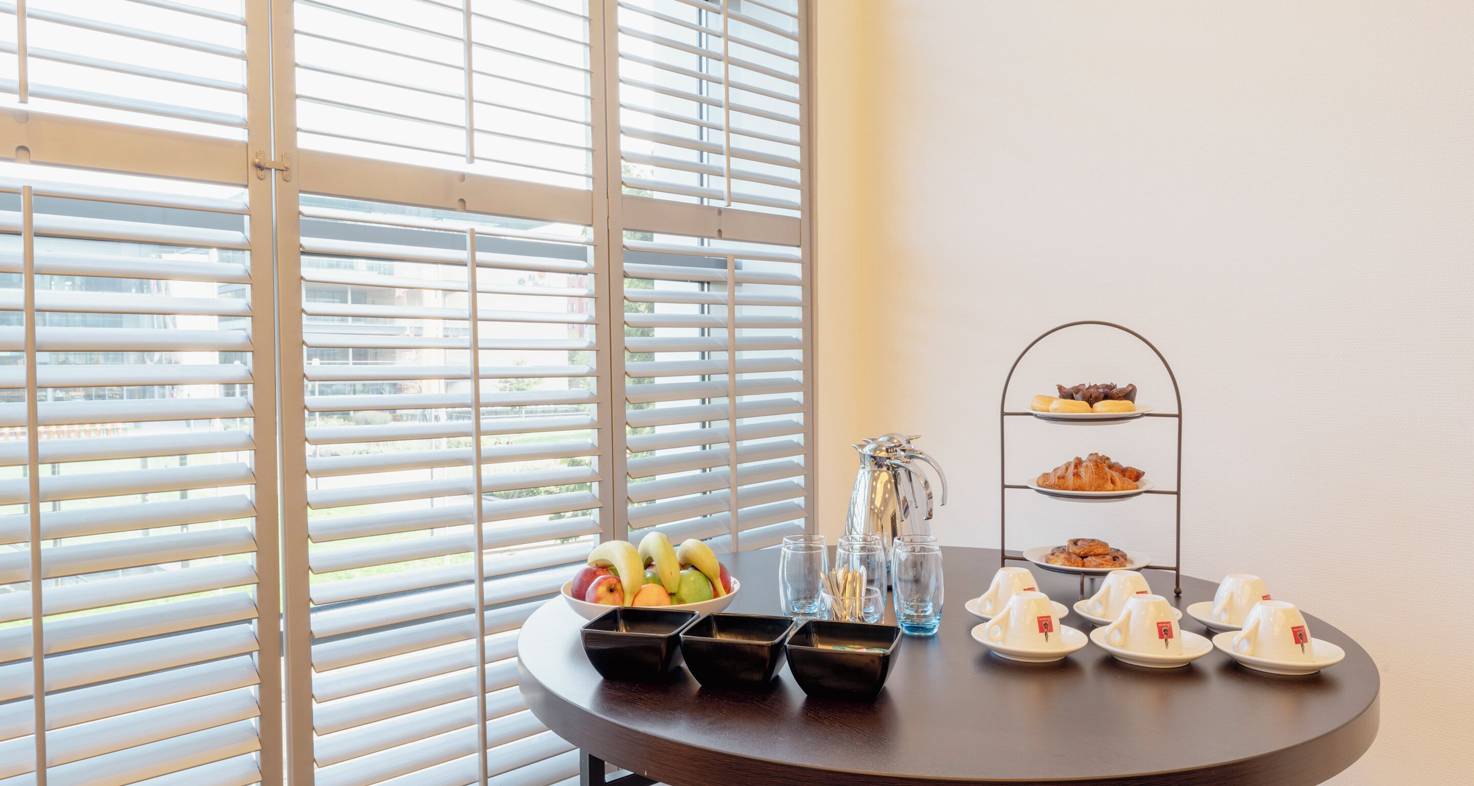A breakfast setup with fruits, pastries, and tea cups on a table near a window with blinds.