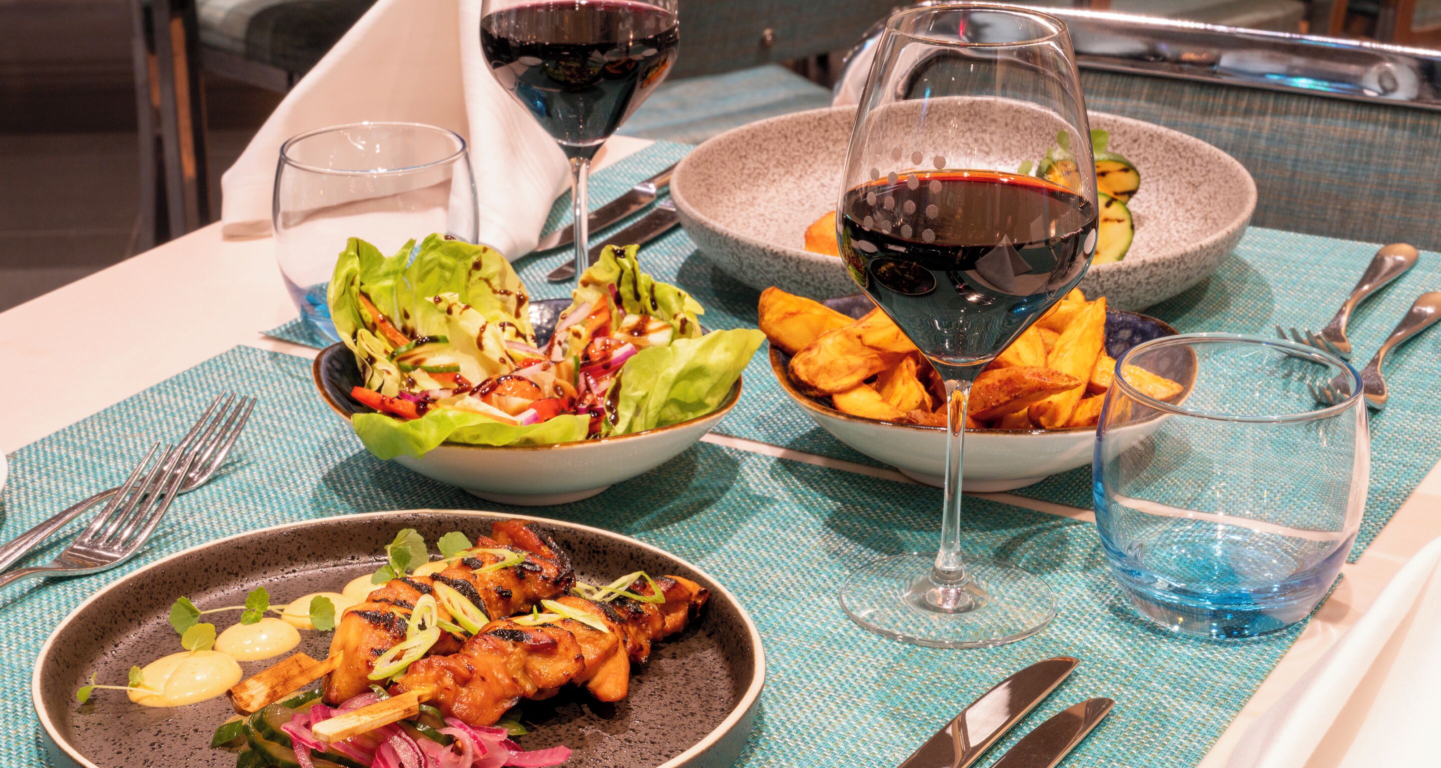 A table set with a salad, grilled chicken, and a bowl of fries, accompanied by wine glasses and cutlery.