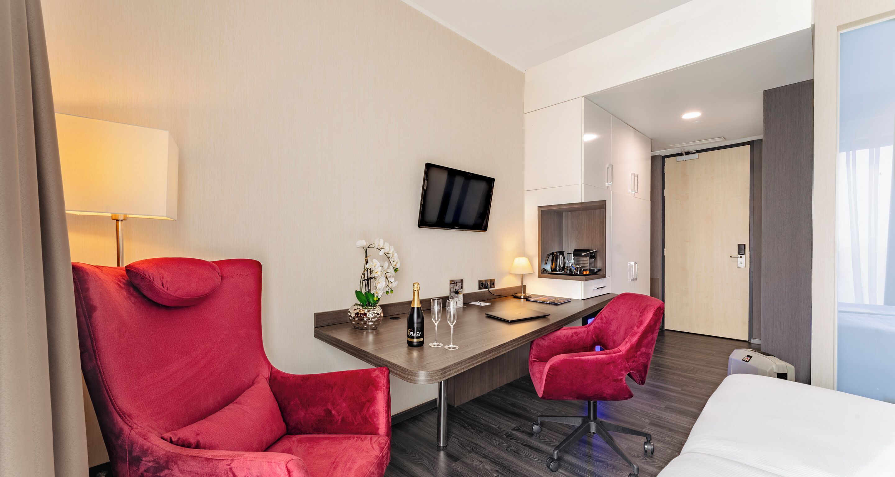 Une chambre d’hôtel moderne avec un grand fauteuil rouge, une petite table sur laquelle se trouvent une bouteille et des verres, un téléviseur fixé au mur et un lit doté d’une tête de lit blanche.