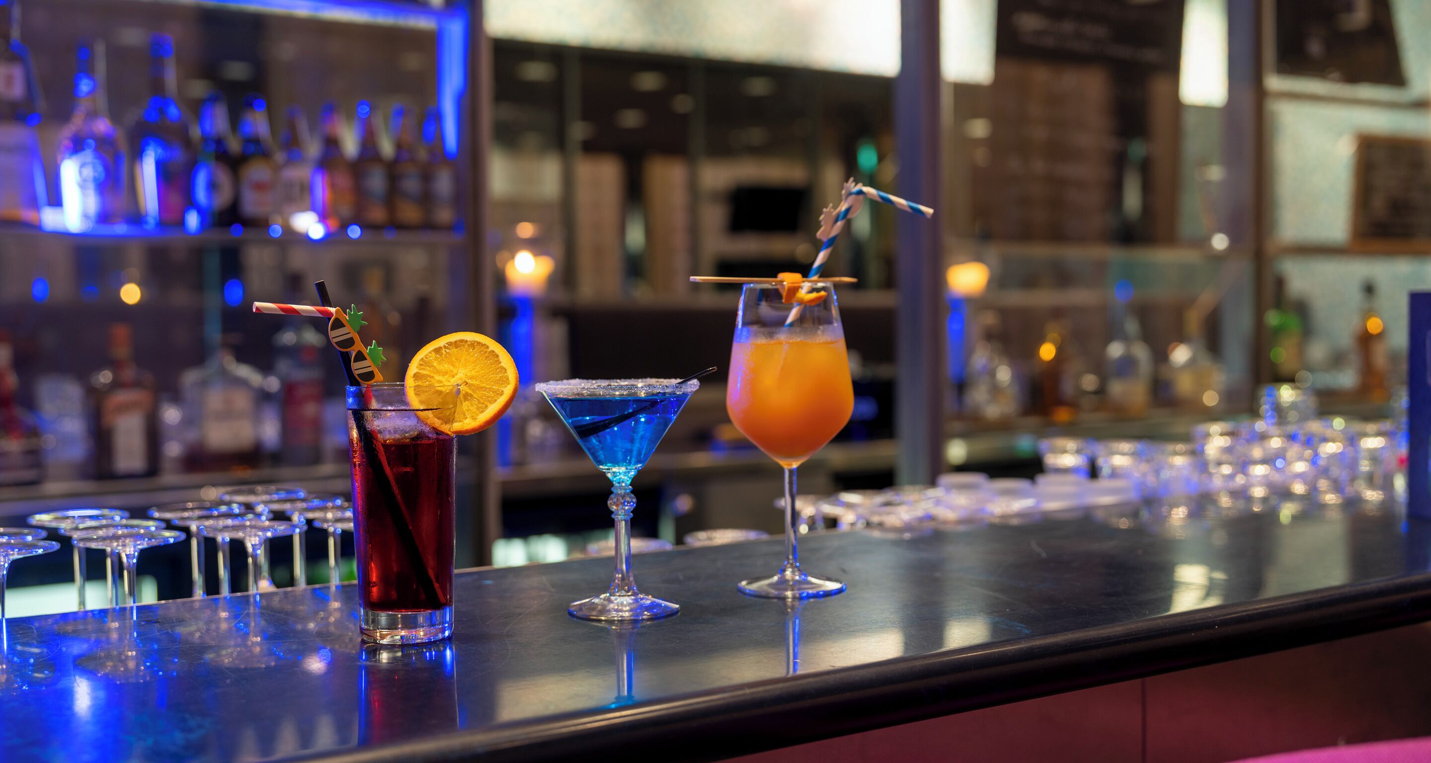 Un bar con tres cócteles, vasos alineados y un fondo borroso de estantes con botellas.