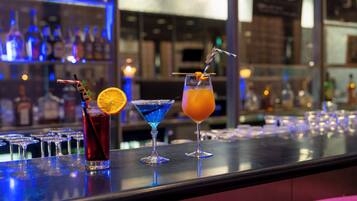 Un bar con tres cócteles, vasos alineados y un fondo borroso de estantes con botellas.