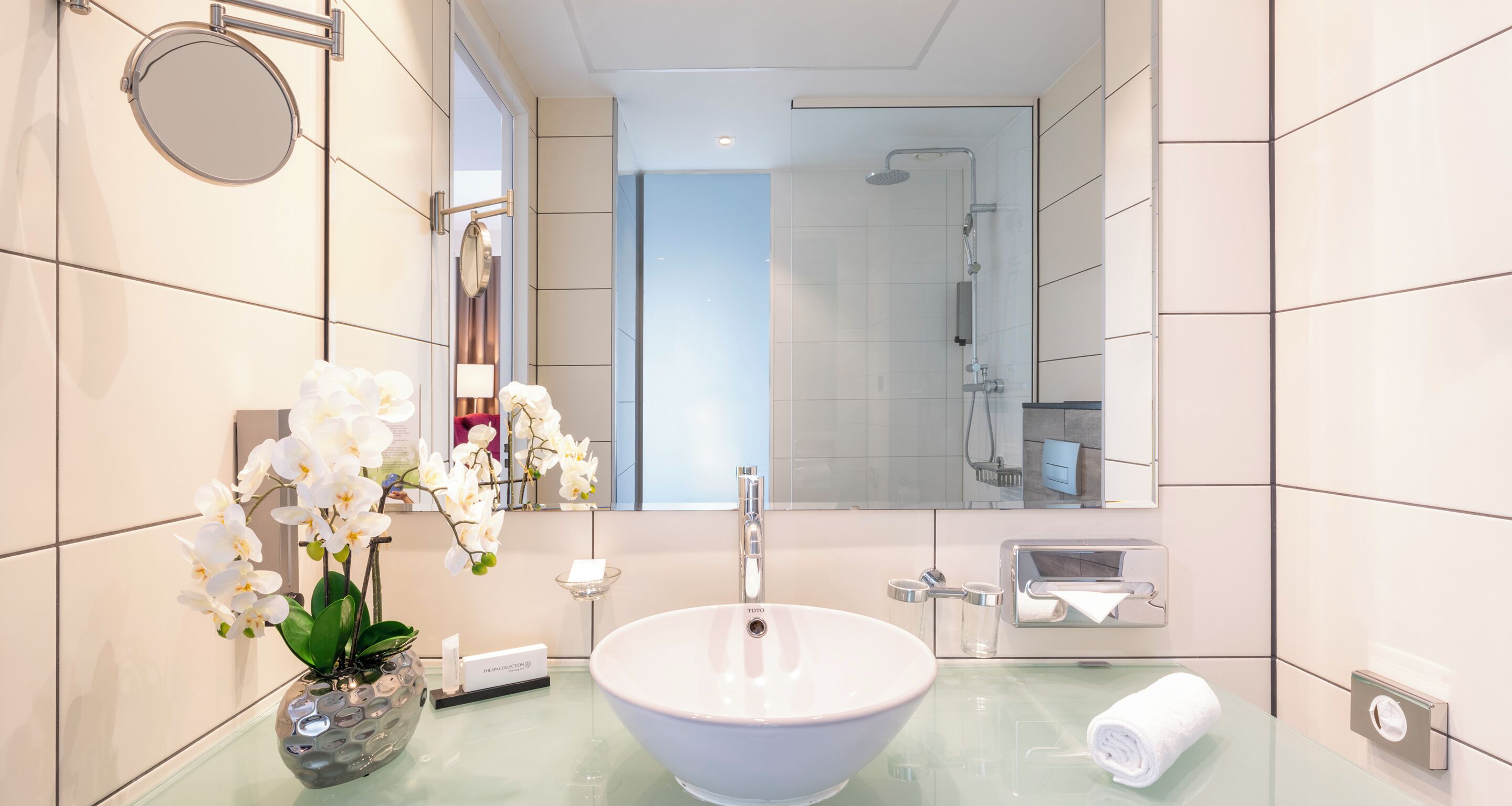A modern bathroom with a large oval sink, a round mirror, a handheld showerhead, and a glass vase with white orchids.