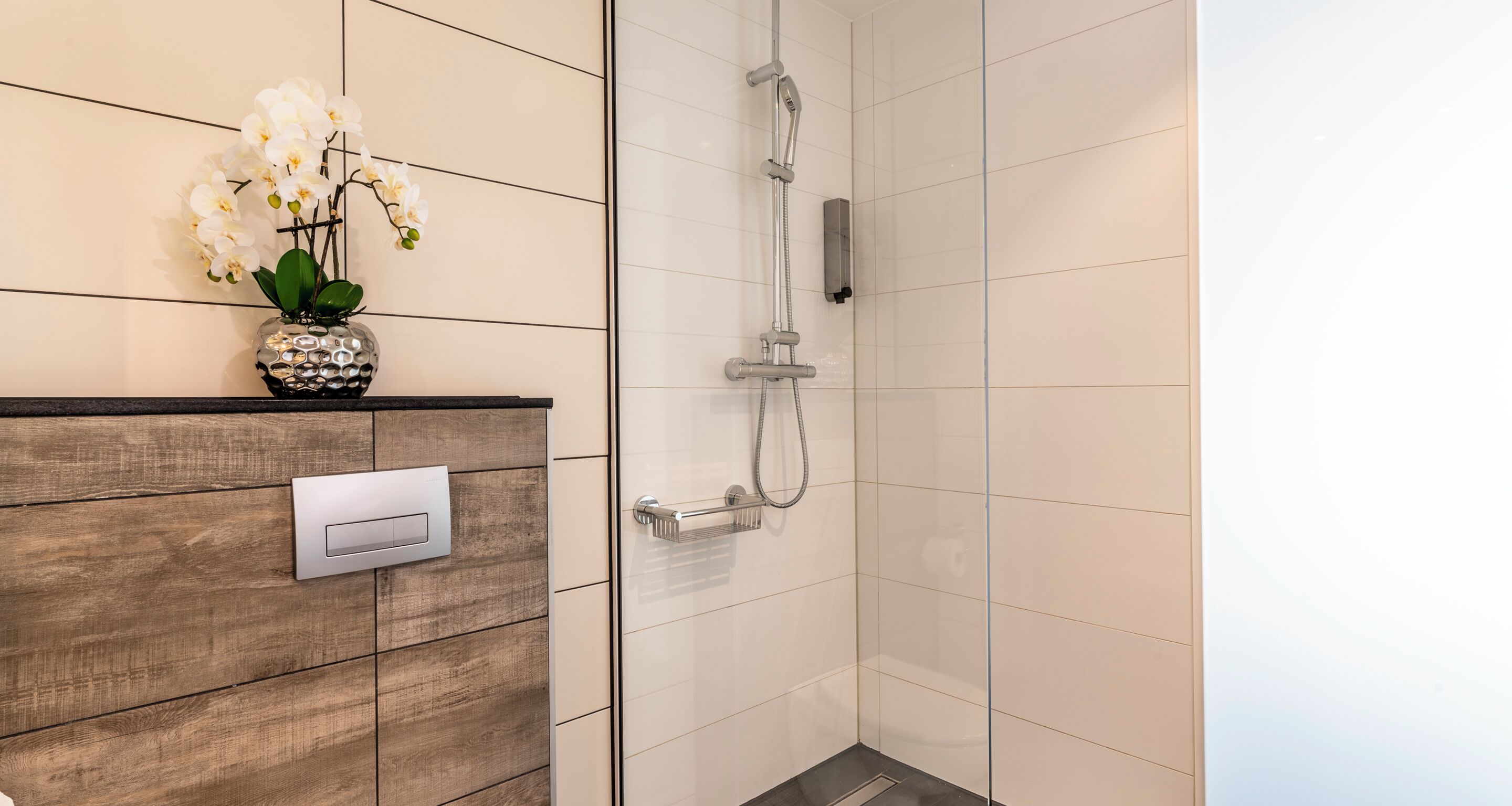 Un baño moderno con un tocador de madera, cabina de ducha de vidrio y un florero con flores blancas sobre el tocador.