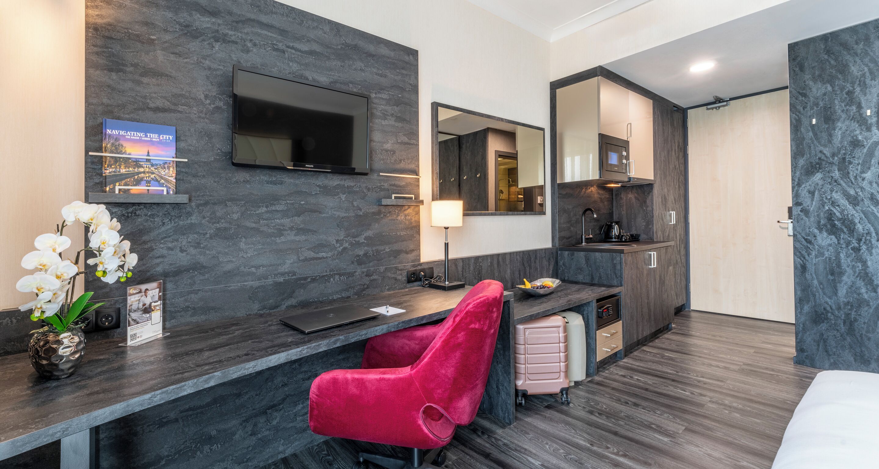 Ein modernes Hotelzimmer mit dunklem Holzschreibtisch, einem roten Sessel, einem Flachbildfernseher, einem Spiegel, einer kleinen Küchenzeile und einem Bett.