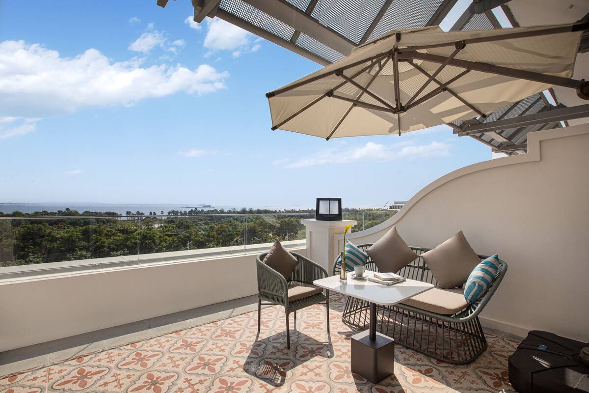 Een balkon met een tafel, stoelen en een parasol. Er is uitzicht op de oceaan en groen in de verte.