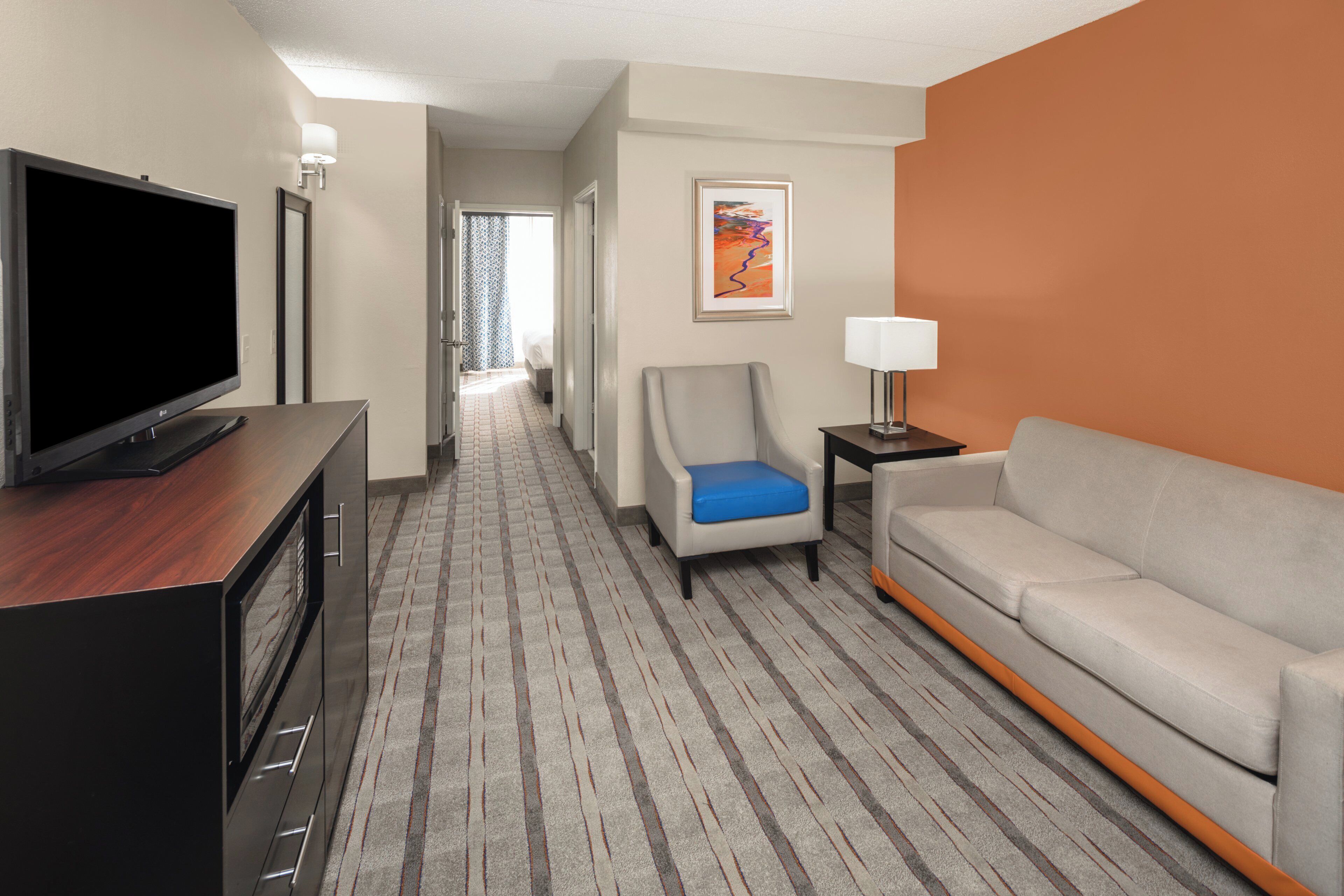 Quarto de hotel com televisão de tela plana, sofá, cadeira, mesa pequena com luminária e corredor que leva a outro quarto.