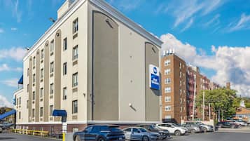 A hotel with a large parking lot, a blue sign indicating Best Western, and a brick building with fire escapes in the background.
