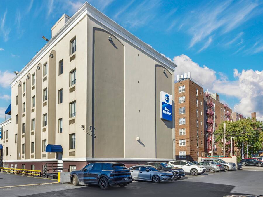 A hotel with a large parking lot, a blue sign indicating Best Western, and a brick building with fire escapes in the background.