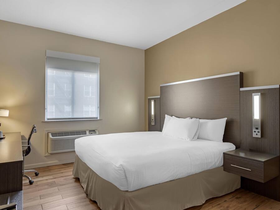 A modern hotel room with a large bed, bedside tables, a desk, and a window with blinds.