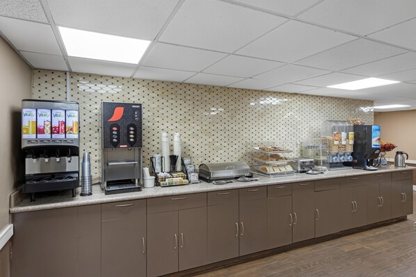 A self-service breakfast area with a coffee machine, a juice dispenser, and a refrigerated display case.