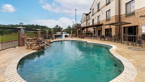 A swimming pool with a stone border, surrounded by a patio area with outdoor seating and a brick building in the background. - Comfort Suites Marshall (Marshall)