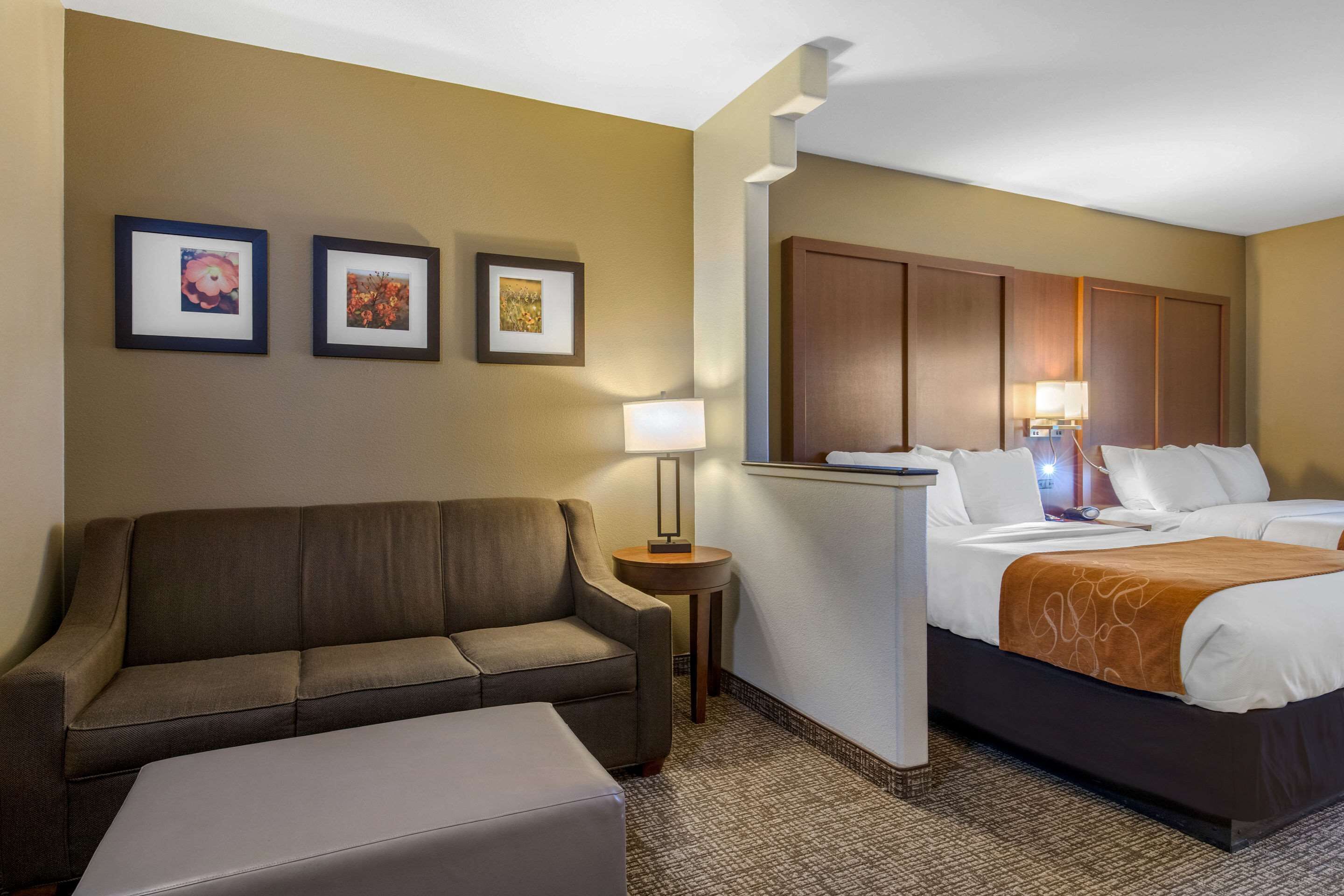A hotel room with a sofa, a bed with a brown blanket, a nightstand with a lamp, and two framed pictures on the wall.