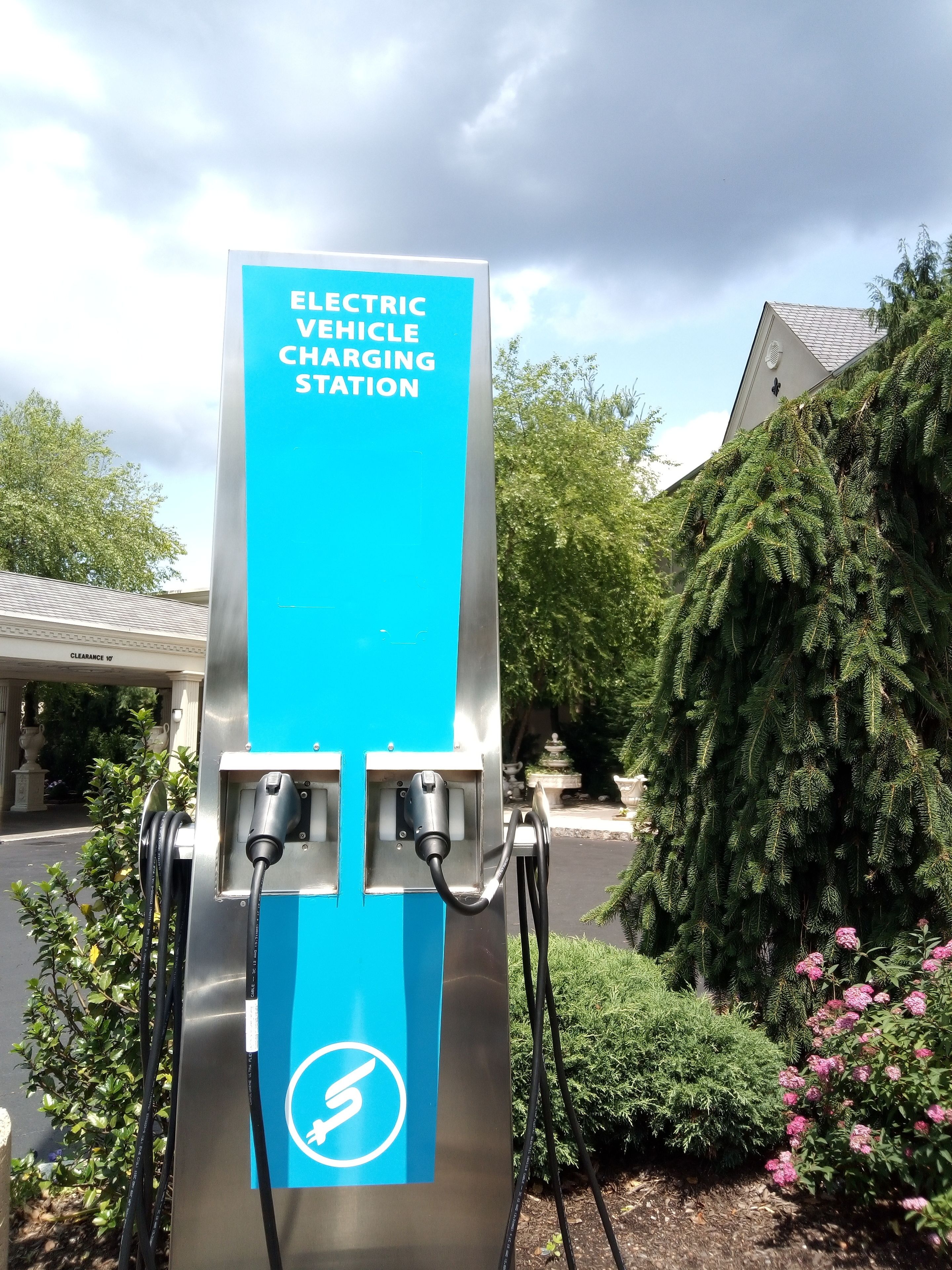 An electric vehicle charging station with a blue sign, surrounded by greenery and a building in the background.
