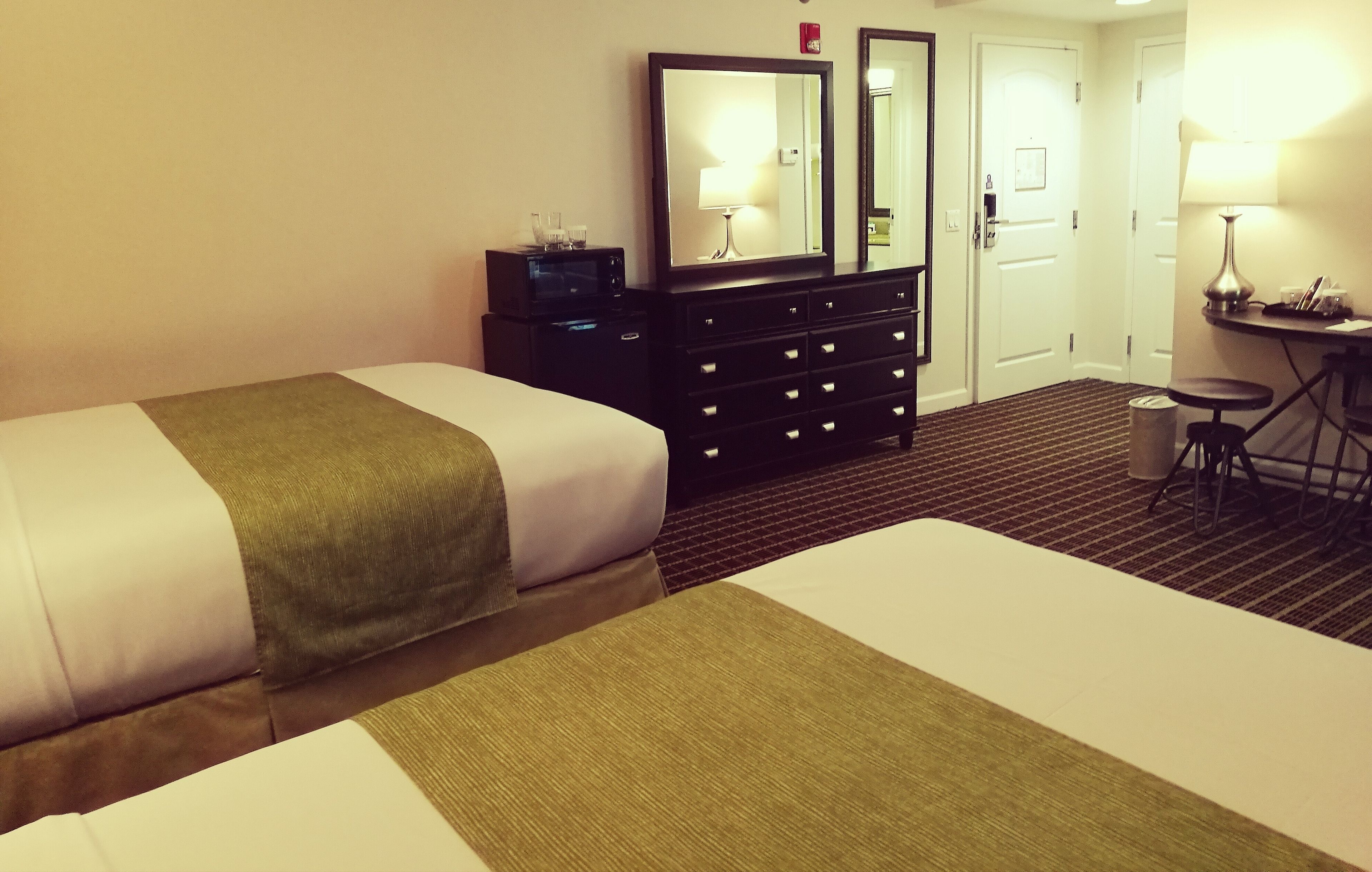 A hotel room with two beds, a dresser with drawers, a mirror, a desk with a lamp, and a trash can.