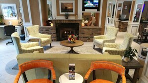 A cozy lounge area with a fireplace, comfortable seating, and a television displaying a news program. - The Wilshire Grand Hotel (West Orange)