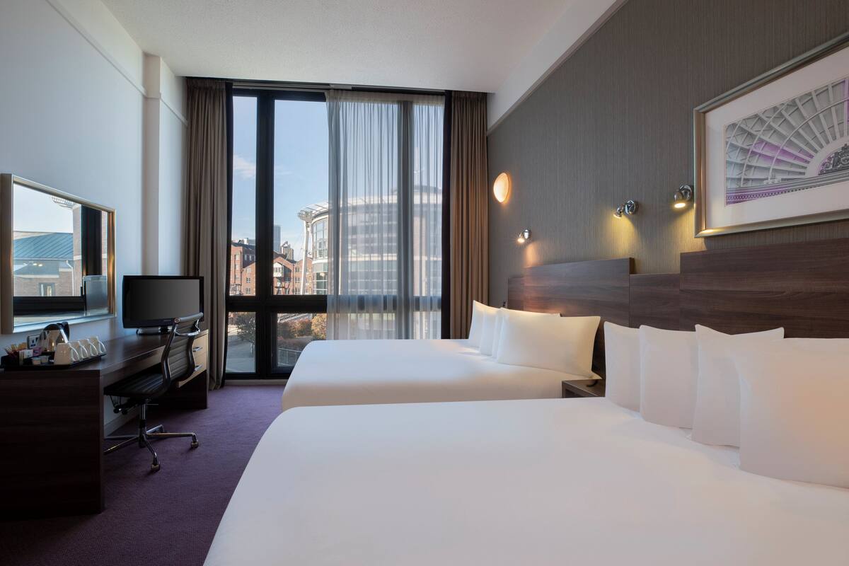 Ein Hotelzimmer mit zwei Betten, einem Schreibtisch mit Computer, einem Fernseher und einem großen Fenster mit Vorhängen.