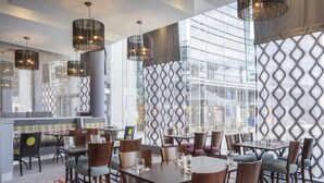 A modern dining area with multiple tables and chairs, large windows with patterned curtains, and hanging pendant lights. - Leonardo Hotel Leeds  (Leeds)