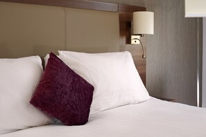 A neatly made bed with white and purple pillows, a wall-mounted lamp, and a wooden headboard. - Leonardo Hotel Leeds  (Leeds)