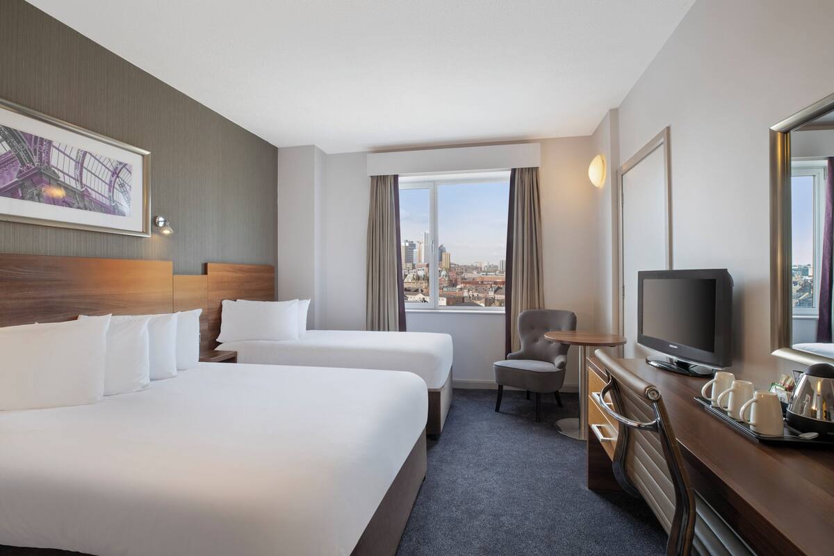 Ein Hotelzimmer mit zwei Betten, einem Schreibtisch mit Fernseher, einem Sessel und einem Fenster mit Blick auf die Stadt.