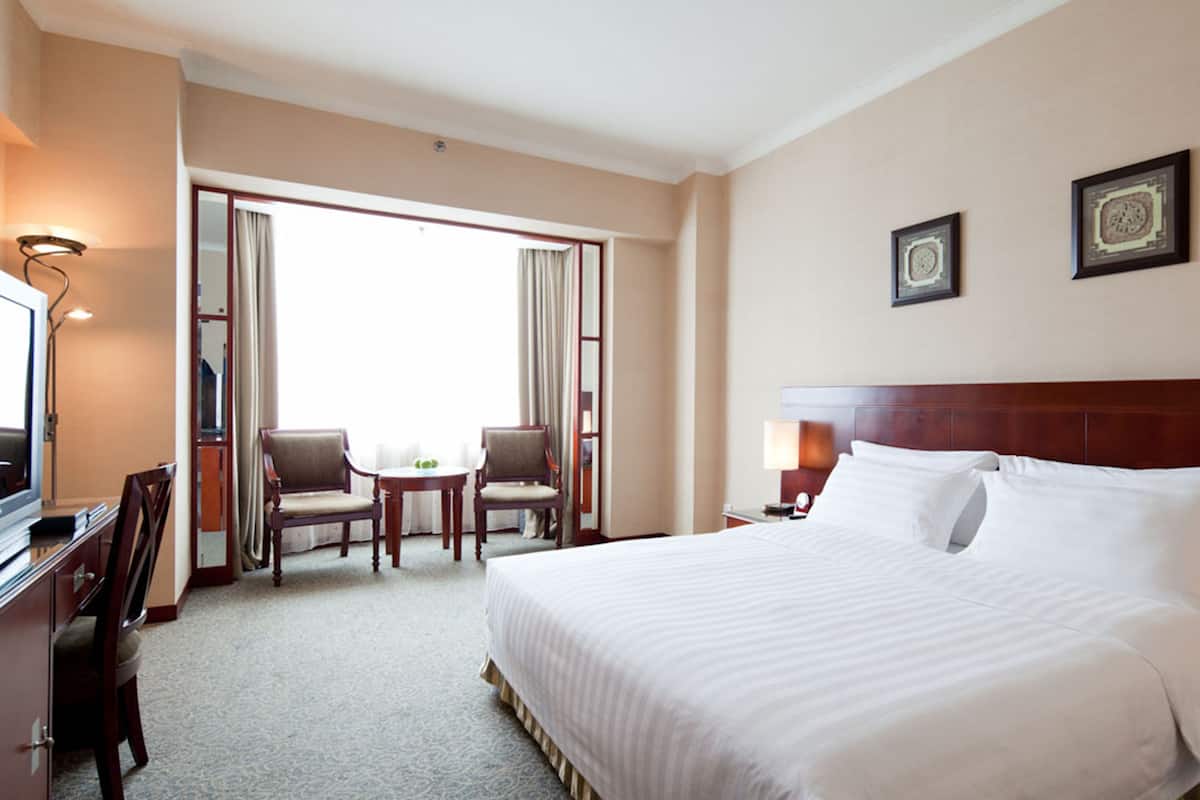 A hotel room with a large bed, a desk with a chair, a small table with two chairs, and a large window with curtains.