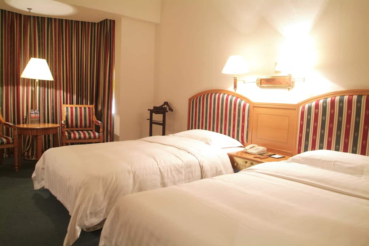 A hotel room with two beds, a small table with a chair, a lamp, and a bedside table with a phone.