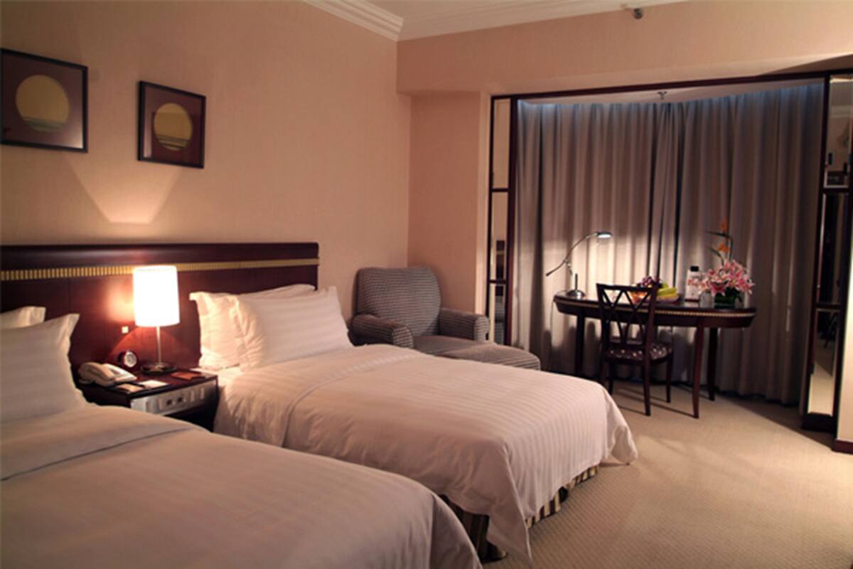 A hotel room with two beds, a desk with a chair, a lamp, and a view of the outside through a curtain.