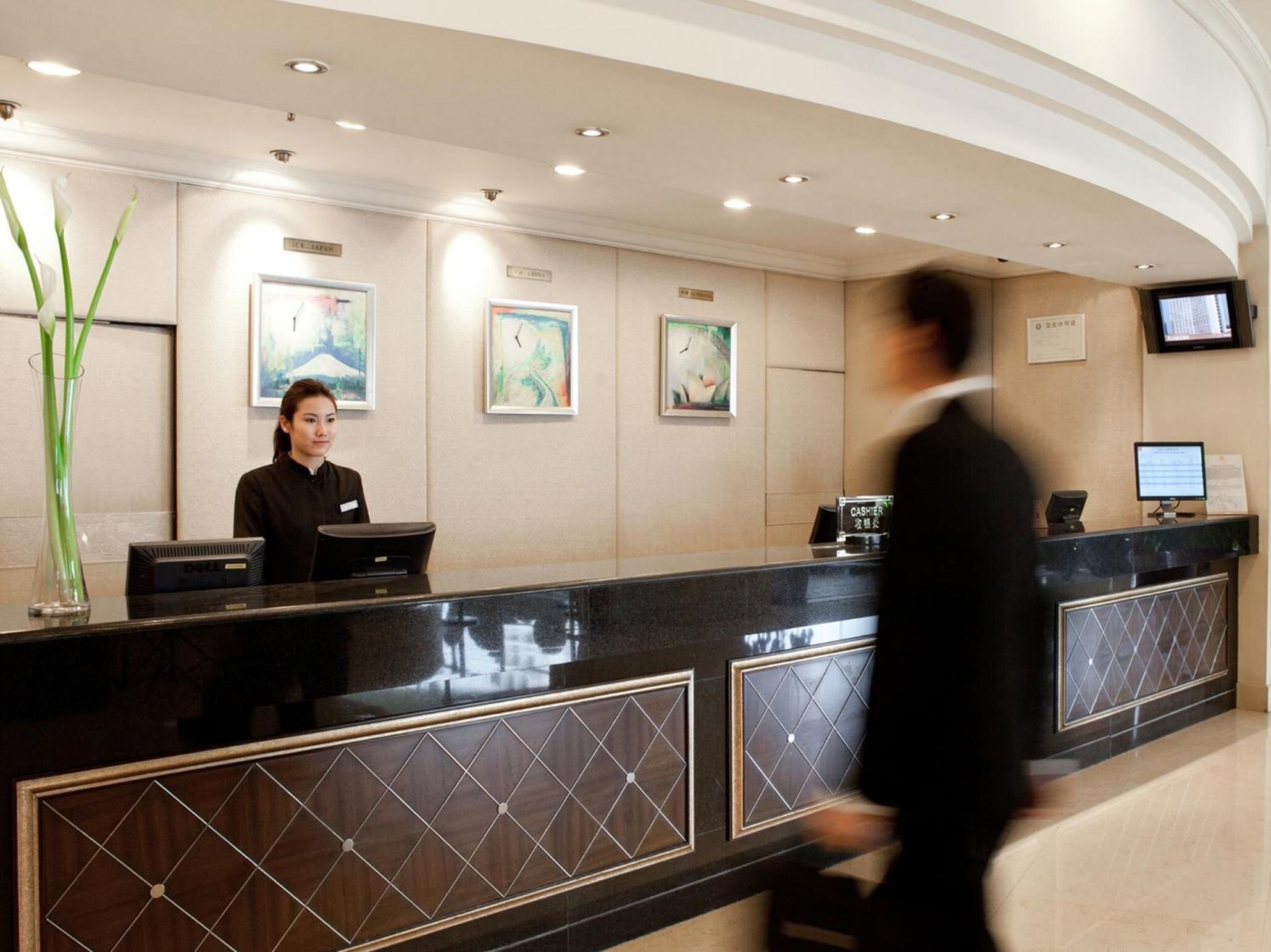 Zona de recepción de un hotel, con un recepcionista, una computadora, una caja registradora y obras de arte enmarcadas en la pared.