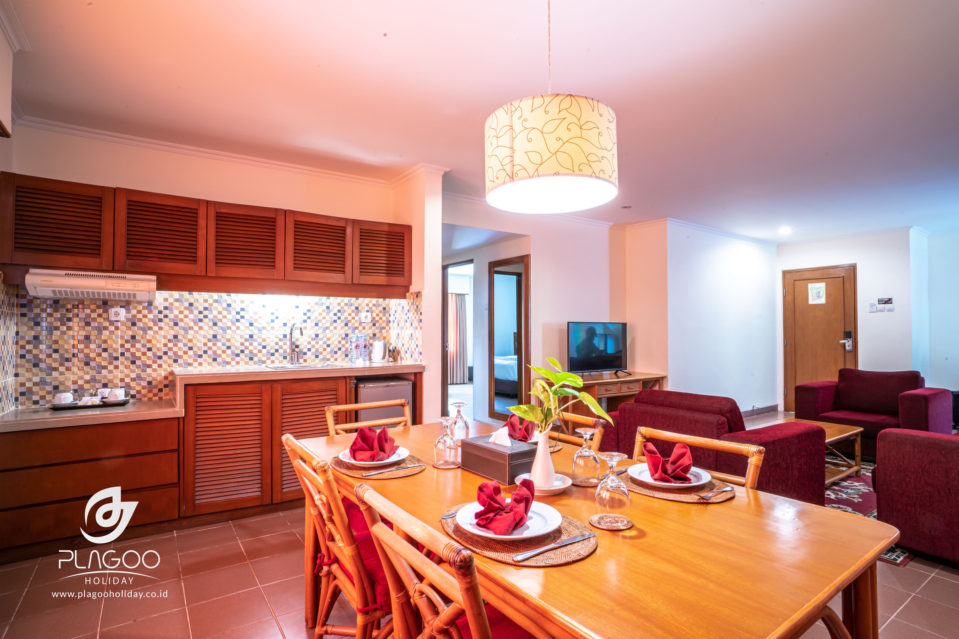 A well-lit dining area with a wooden table set for four, a television in the background, and a red sofa in the living area.