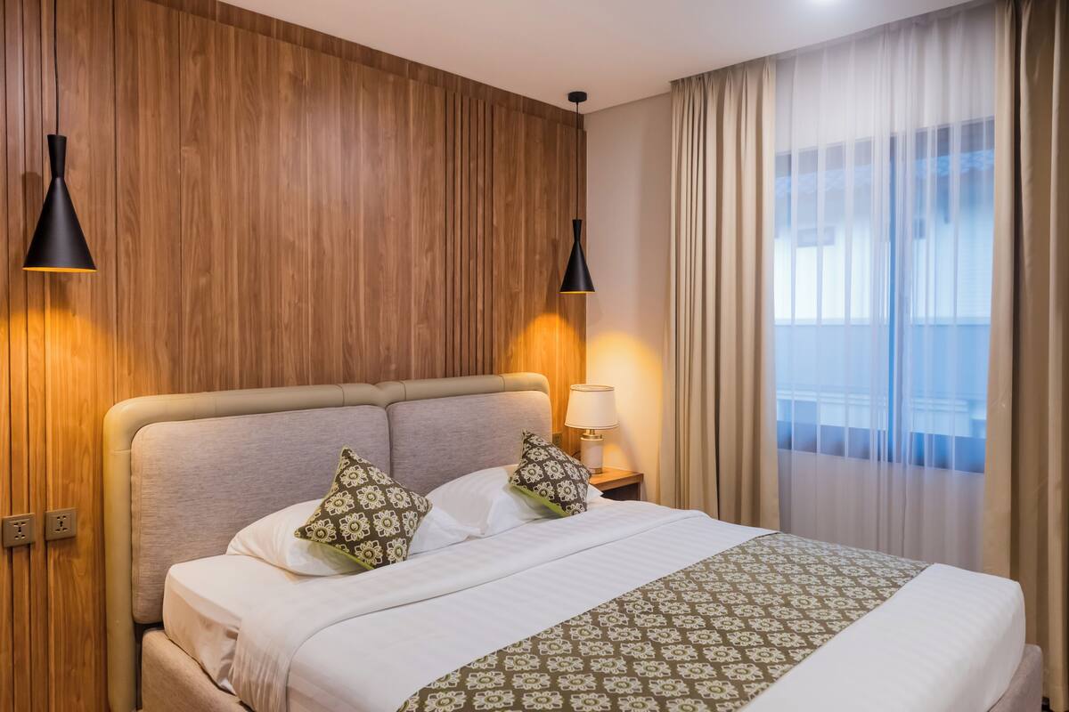 Une chambre d’hôtel avec un mur lambrissé en bois, un lit avec une literie à motifs, deux lampes de chevet et une vue à travers des rideaux transparents.