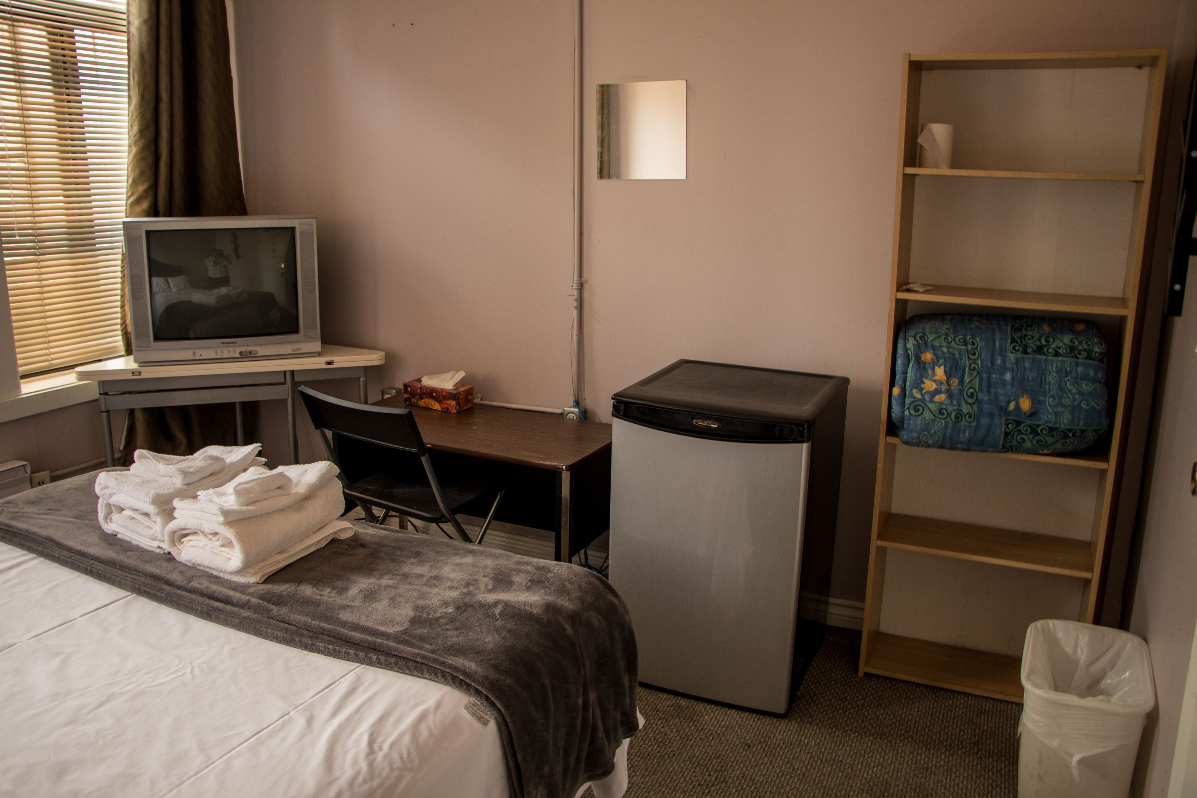 Une chambre d’hôtel comprenant un lit, un bureau avec une télévision, un petit réfrigérateur, une chaise, une étagère avec un objet décoratif et une poubelle.