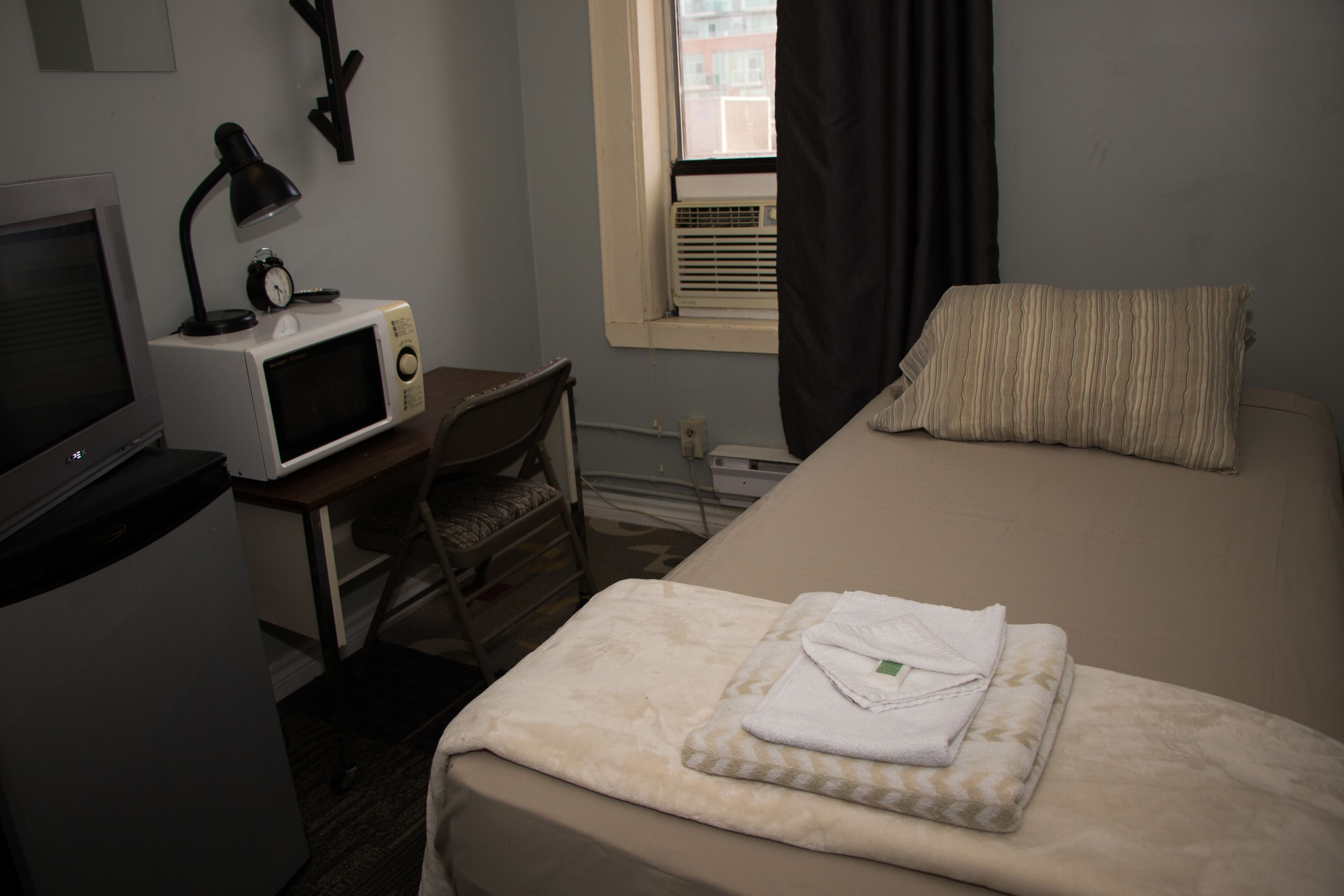 A compact hotel room with a bed, a desk with a computer and a lamp, a microwave, and a window with curtains.