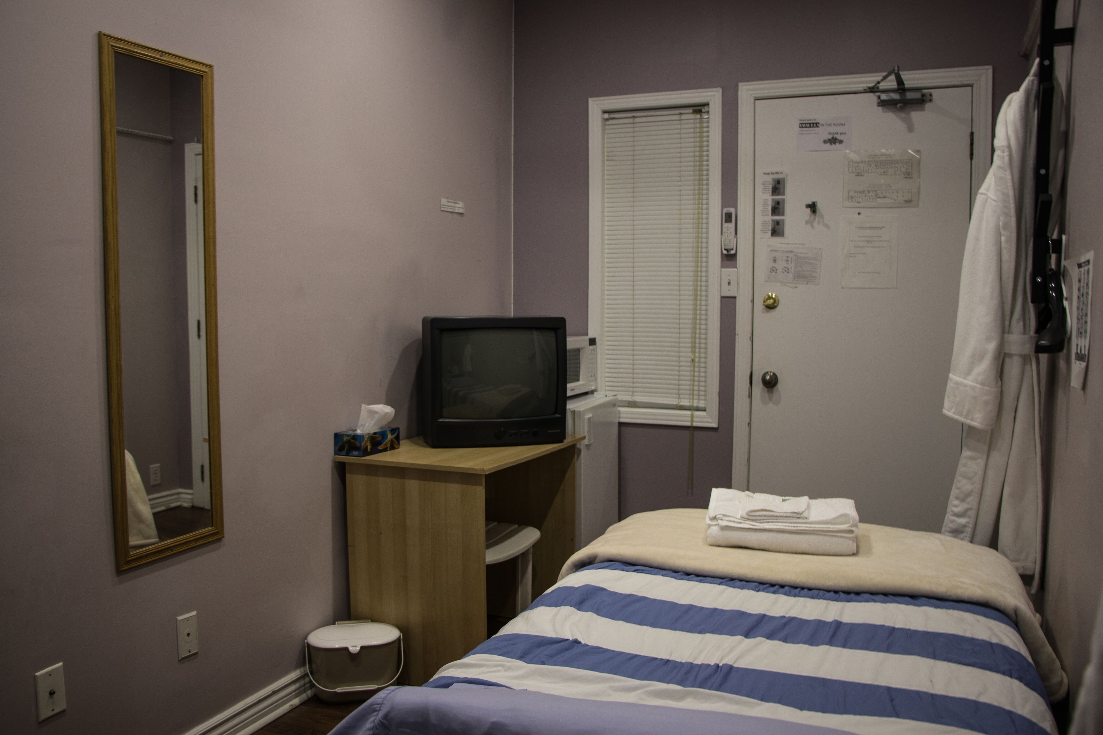 A hotel room with a bed, a television on a wooden desk, a mirror, a door with a window, and a towel rack.