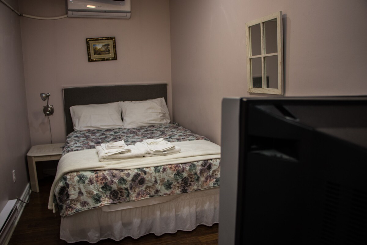 Een netjes opgemaakt bed met een bloemenbedekking en witte kussens, een bedlampje, een klein tafeltje en een foto aan de muur.