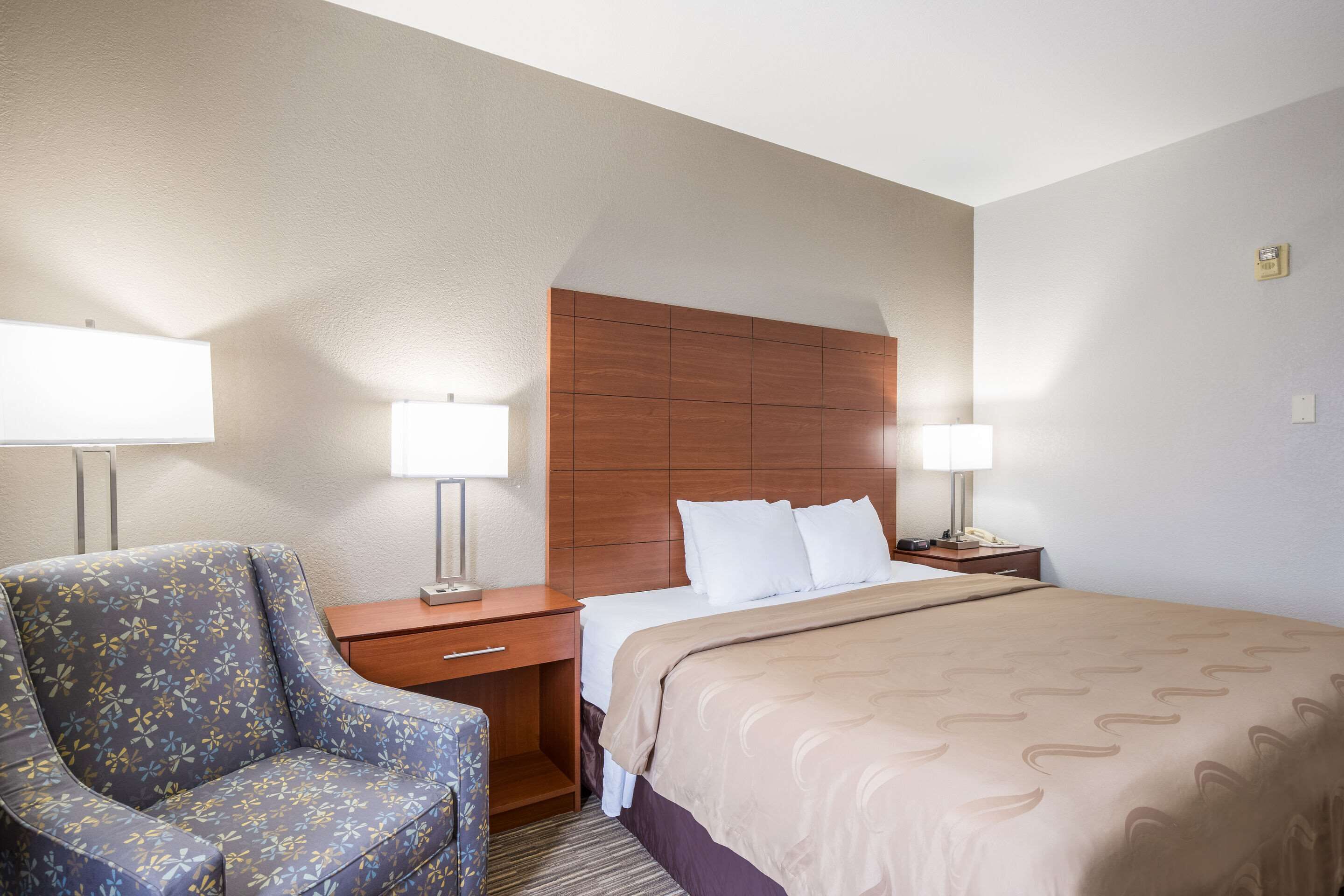 A hotel room with a bed, a nightstand, a lamp, a chair, and a wooden headboard.