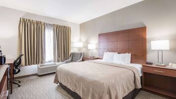 A hotel room with a large bed, a desk with a chair, a bedside table with a lamp, and a window with curtains.