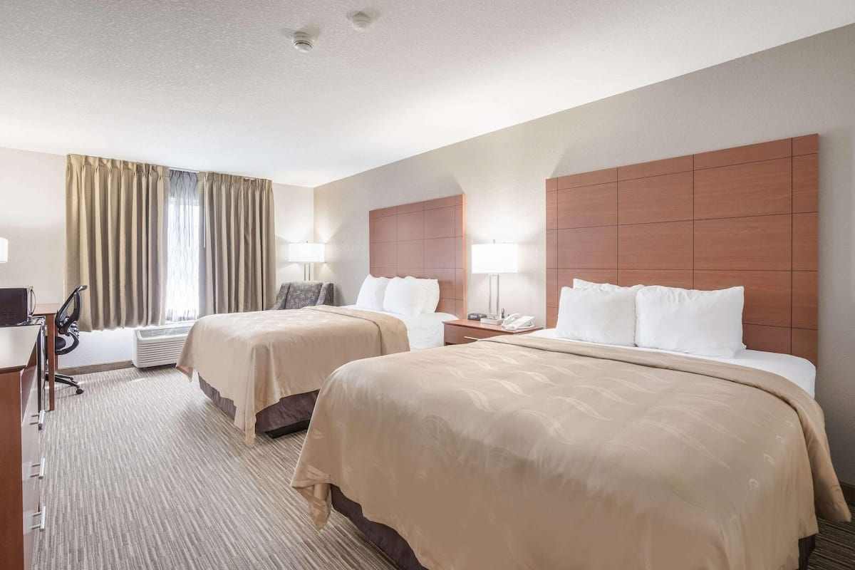 Une chambre d’hôtel avec deux lits, un bureau, une chaise, une lampe et une fenêtre avec des rideaux.