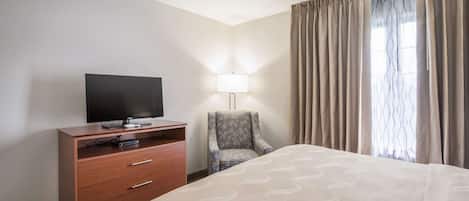 Ein Hotelzimmer mit einem Bett, einem Fernseher auf einer Holzkommode, einem Sessel und einem Fenster mit Vorhängen.