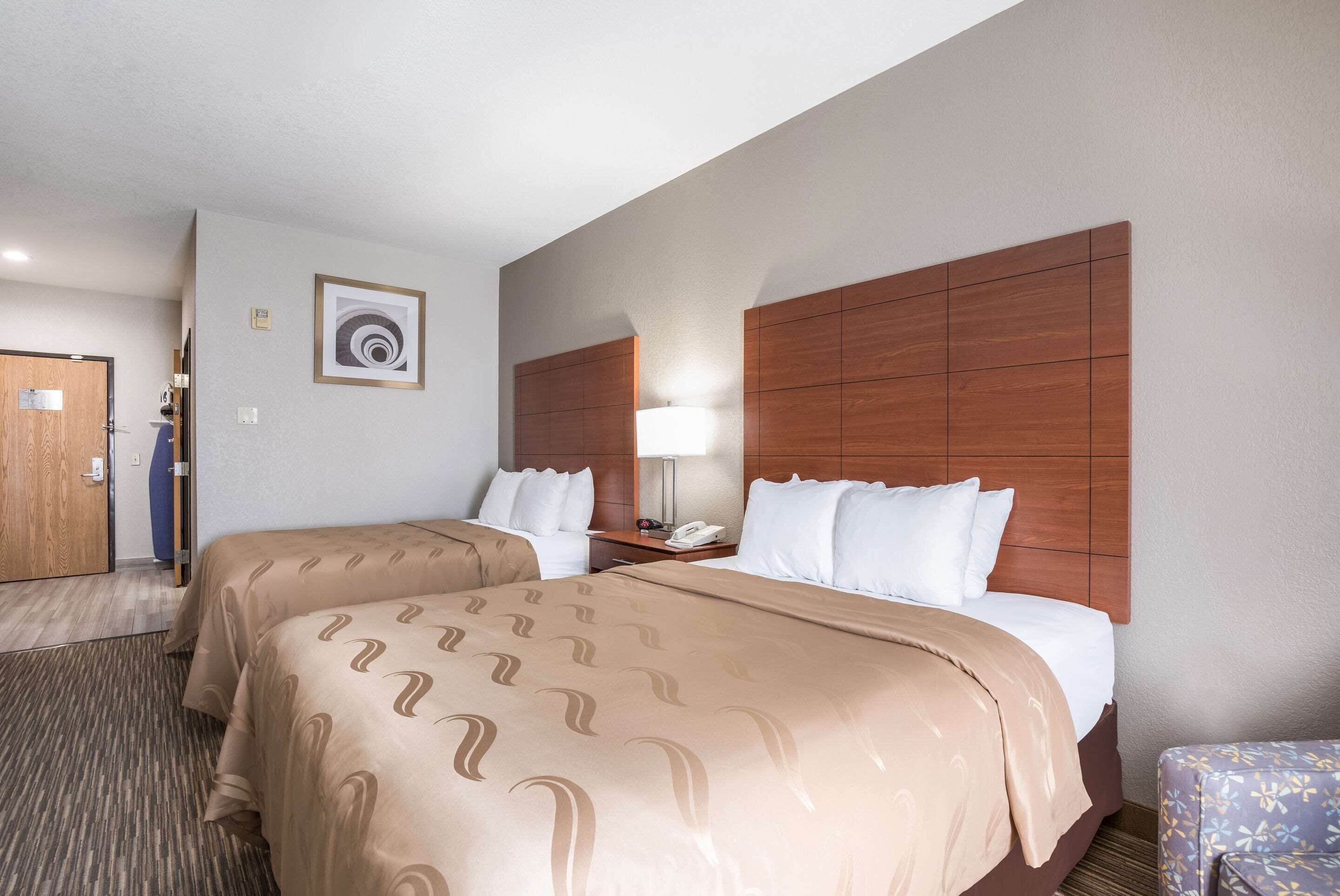 A hotel room with two beds, a wooden headboard, a bedside table with a lamp, and a framed picture on the wall.