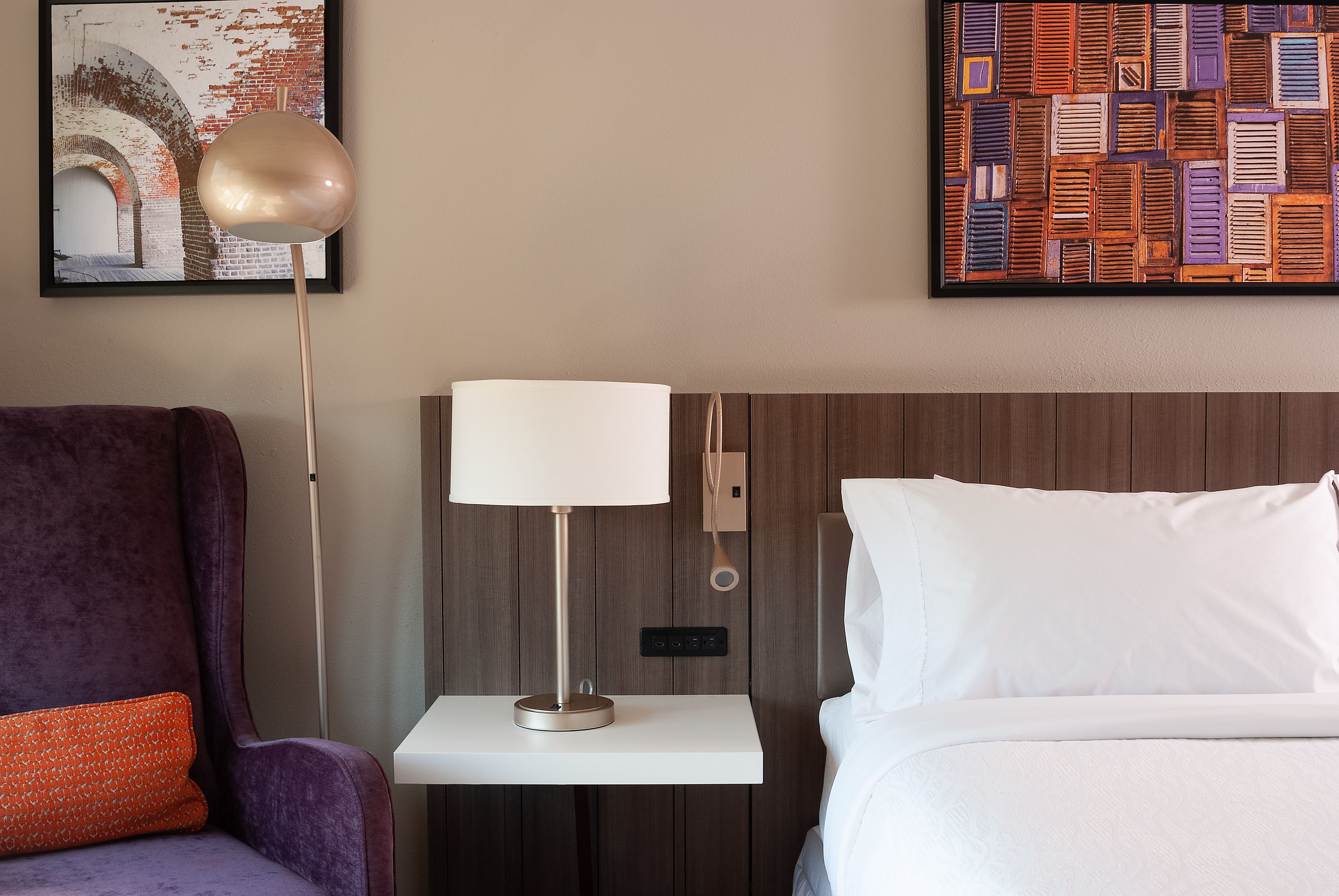 A hotel room with a bed, a nightstand with a lamp, a purple armchair, and a framed artwork on the wall.
