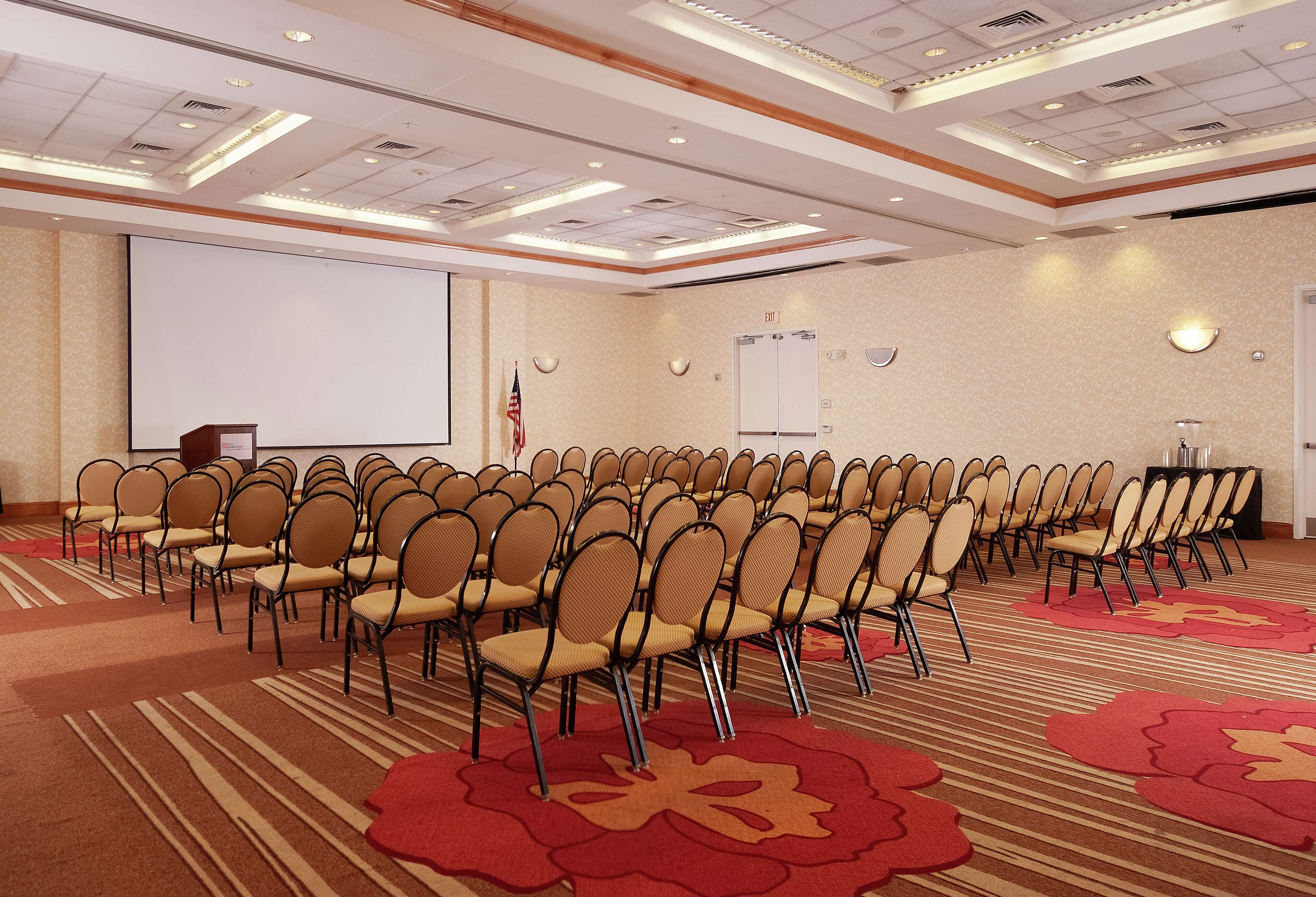 A conference room with rows of chairs, a podium, a projector screen, and an exit sign.