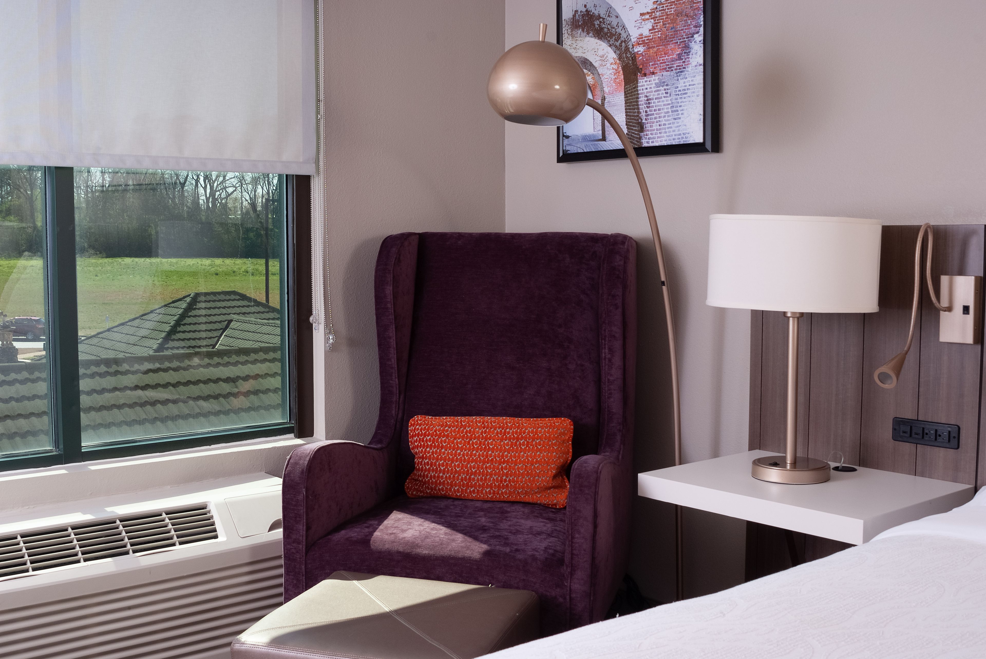 A modern hotel room with a large window, a purple armchair with a red pillow, a bedside table with a lamp, and a framed picture on the wall.