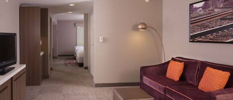 A modern hotel room with a flat-screen TV, a purple sofa with orange pillows, a standing lamp, and a large framed picture on the wall.