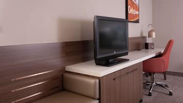 A hotel room with a wooden bench, a desk with a flat-screen TV, a red chair, and a lamp.