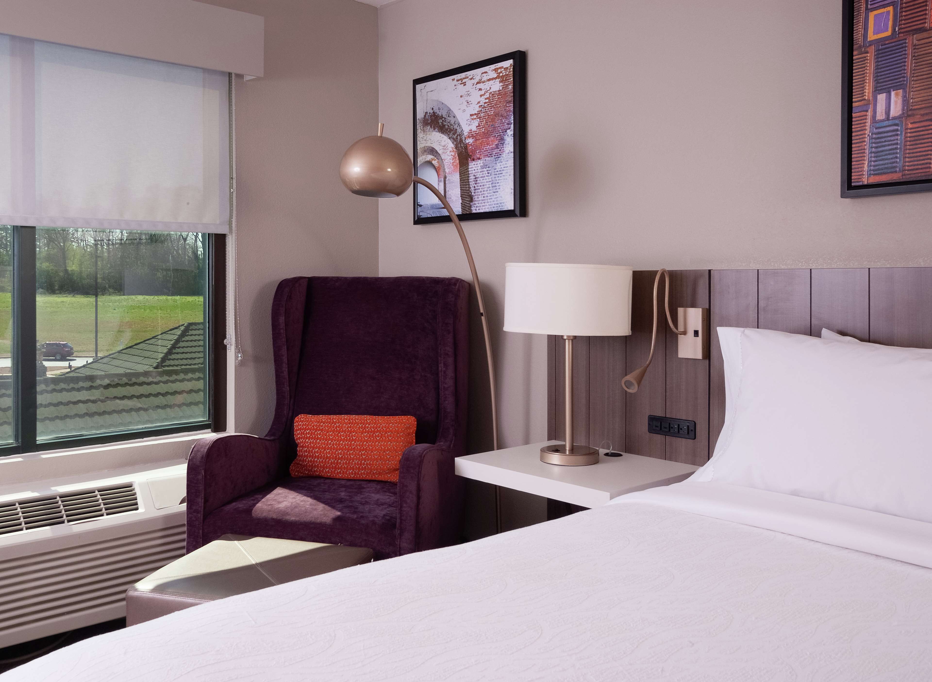 A hotel room with a large bed, a chair with a red cushion, a bedside table with a lamp, and a window with a view of a green landscape.