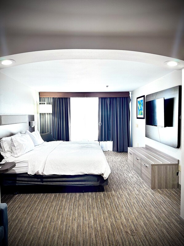 Ein modernes Hotelzimmer mit einem großen Bett, einem Flachbildfernseher, einer hölzernen Kommode und einem großen Fenster mit Vorhängen.